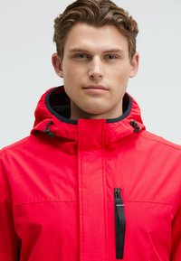 Jeune homme avec des cheveux bruns courts portant une veste à capuche rouge vif avec une poche zippée sur la poitrine et des cordons réglables pour la capuche.