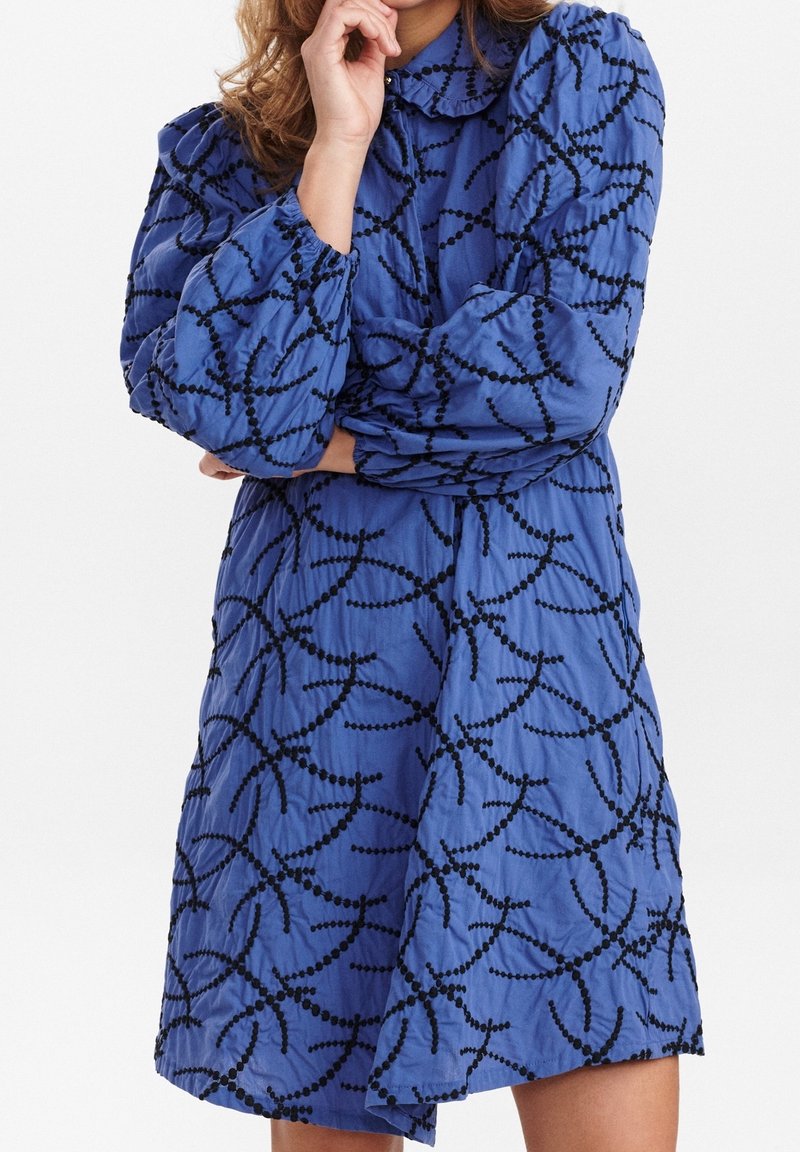 Robe bleue avec un col à revers, des manches bouffantes et des motifs tourbillonnants en perles noires. Longueur courte avec un tissu texturé.