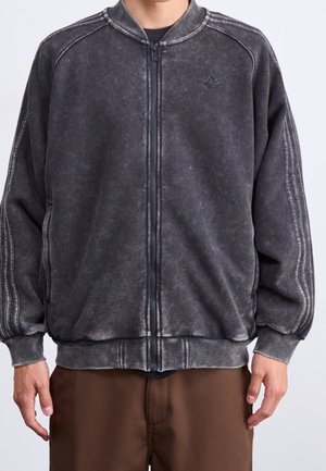 Homme portant une veste zippée noire délavée avec des poignets côtelés et un pantalon marron, visible du cou aux hanches sur un fond uni.