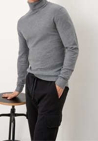 Pull à col roulé gris en tissu doux, avec manches longues et poignets côtelés, assorti à un pantalon cargo noir et un tabouret en bois.