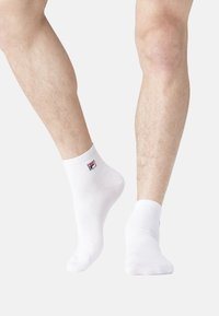 Chaussettes blanches en coton jusqu'à la cheville avec un revers côtelé, présentant un petit logo coloré de la marque sur le côté. Texture lisse et design ajusté.