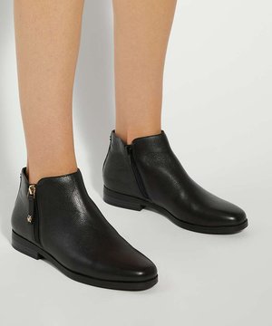 Bottes en cuir noir montant à la cheville avec fermeture éclair latérale, portées sur des jambes nues sur un fond clair uni.