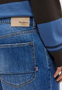 Μπλε τζιν από τζιν ύφασμα με δερμάτινη ετικέτα με την ένδειξη "Pepe Jeans London" στη μέση, με εμφανείς ραφές και μια μικρή βρετανική σημαία στην τσέπη.