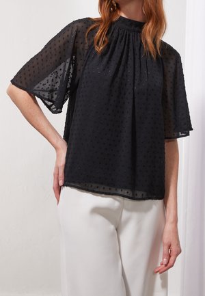 Vrouw die een doorschijnende zwarte blouse met getextureerde stippen en losse mouwen draagt, gecombineerd met een witte broek, poseert met één hand op de heup.