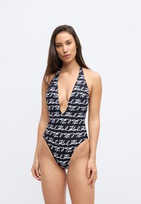 Μαγιό με βαθύ ντεκολτέ σε σχήμα V, κατασκευασμένο από ελαστικό ύφασμα. Διαθέτει μαύρο φόντο με μοτίβο άσπρων χειρόγραφων κειμένων και ψηλή κοπή στα πόδια.