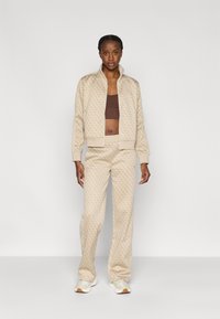 Μπεζ φόρμα με μοτίβο, με ζακέτα με φερμουάρ και αντίστοιχο παντελόνι, με ριμπ μανσέτες και καφέ εσωτερική μπλούζα. Λευκά αθλητικά παπούτσια ολοκληρώνουν το ντύσιμο.