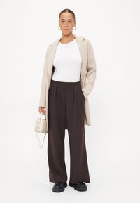 Beige ullblandad kappa över en vit anpassad topp, tillsammans med högmidjade bruna veckade byxor och svarta kraftiga skor. Håller en liten beige handväska.