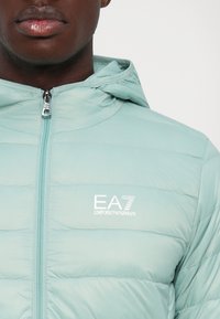 Bărbat îmbrăcat cu o jachetă tip quilted de culoare verde deschis, cu glugă, logoul EA7 Emporio Armani pe pieptul stâng, fermoar parțial închis.