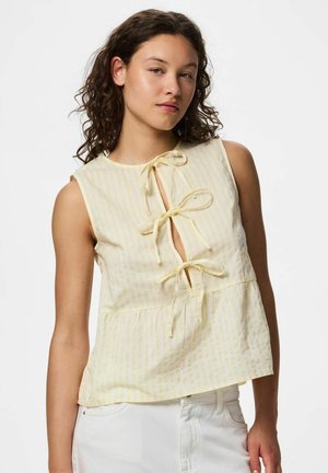 Młoda kobieta o kręconych włosach, ubrana w żółty, bezrękawnikowy top w paski z trzema wiązanymi kokardkami z przodu oraz białe spodnie.