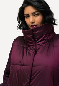 Lila Puffy-Jacke mit hohem Kragen, ausgestattet mit einem gesteppten Design und glänzender Textur. Das Akzentmaterial an den Ärmeln ist aus Spitz und strukturiert.