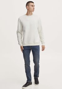 Lichtcrème sweatshirt met geribbelde manchetten en zoom, gecombineerd met donkerblauwe jeans en zwarte lage sneakers, staande tegen een neutrale achtergrond.