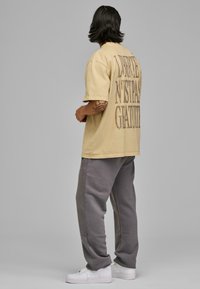 Tricou oversized bej cu text imprimat pe spate, asociat cu pantaloni de trening gri și adidași albi. Material neted și croială lejeră.