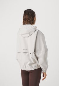 Helle graue wasserdichte Jacke mit Kapuze, elastischen Bündchen und Raffung am Saum. Verfügt über einen vertikalen reflektierenden Streifen auf dem Rücken.