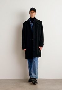 Manteau noir avec col, à boutons sur le devant, porté par-dessus une chemise en denim et un col roulé. Jean bleu à jambes larges, casquette noire et chaussures noires.