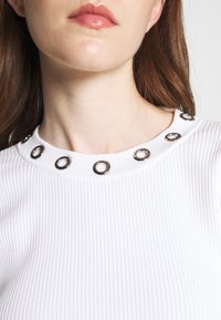 Haut blanc à côtes avec un col rond orné d'accents circulaires en argent. Texture lisse avec une coupe ajustée.