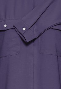 Vijolična srajca z gladko teksturo, ki ima gumbaste manšete, dve prsni žepu in bele gumbe. Čist, minimalističen dizajn.