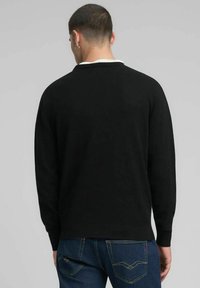 Pull en maille noire avec col blanc, manches longues et poignets côtelés. Porté avec un jean bleu présentant un design de poche cousue.