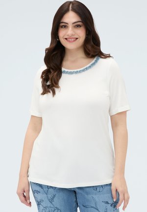 Donna con lunghi capelli castani sorridente, che indossa una maglietta bianca a maniche corte con decorazioni blu intorno allo scollo e jeans blu con fantasia.