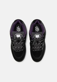 Adidași negri și gri cu un model texturat în carouri. Prezintă accente de piele întoarsă, șireturi negre și un guler căptușit de culoare mov. Logo-ul Vans este afișat.