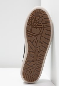 Semelle de chaussure noire avec une semelle en caoutchouc brun texturé présentant un motif géométrique et une semelle intermédiaire de couleur claire. Logo de la marque visible.