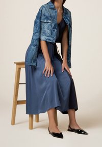 Jeansjacke mit Blumenmuster, marineblaues Satinkleid und schwarze spitze Ballerinas. Ein Holzstuhl bietet eine sitzende Pose, die die Details der Kleidung betont.