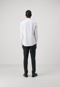 White long-sleeve shirt with a classic collar, paired with dark green trousers and brown shoes, viewed from the back. Smooth texture, minimal design.