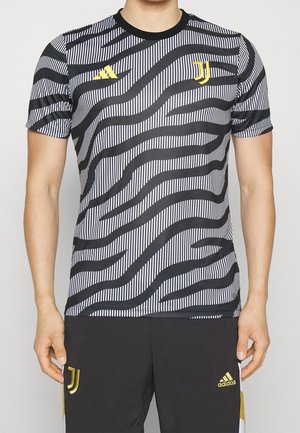 Mand iført sort- og hvidstribet Juventus fodboldtrøje og sorte Adidas-shorts med gule logoer, stående mod en ensfarvet baggrund.