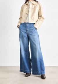 Beżowa krótka kurtka, jasnoszary top, niebieskie dżinsy z szerokimi nogawkami, brązowe botki na obcasie; gładka tekstura, jednolite kolory i swobodny fason.