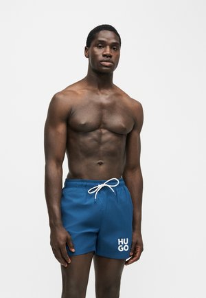 Homme musclé se tient face à l’avant, portant un short de bain bleu Hugo avec un cordon blanc sur un fond clair uni.