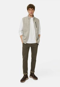 Graue Fleece-Weste über einem weißen Langarmshirt, olivgrüne Hose, braune Wanderschuhe. Enthält einen vorderen Reißverschluss und Seitentaschen.