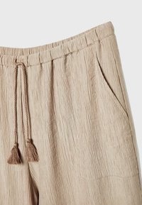 Pantalon beige texturé avec taille élastique et cordons à pompons, doté d'une poche latérale droite.