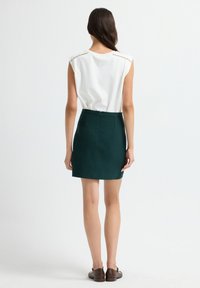 Top bianco senza maniche con decorazioni sulle spalle, abbinato a una gonna verde aderente. L'outfit presenta una cerniera sul retro e una texture liscia.