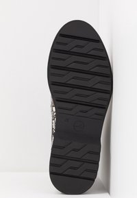 Semelle d'une chaussure noire avec motif de crampons, marquage de pointure 37, et logo circulaire, affichée verticalement sur fond blanc.