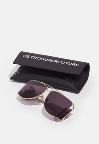 Occhiali da sole con una montatura in metallo dorato e lenti viola scuro, esposti insieme a un astuccio nero con la scritta "RETROSUPERFUTURE."