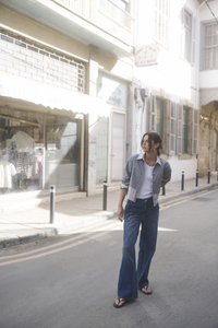 Prugasta crop jakna, bijela majica i široke plave traperice. Scena prikazuje kameniti uličicu i izloge trgovina u mekom suncu. Nošene su japanke.