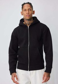 Homme avec des tatouages sur le cou et la main, portant un sweat à capuche noir zippé et un pantalon blanc, debout devant un fond gris clair uni.