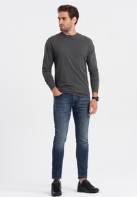 Donkergrijze longsleeve shirt, slim-fit blauwe jeans en zwarte sneakers. Model staat rechtop, met handen ontspannen langs de zij, op een effen achtergrond.
