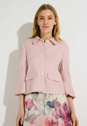 Vrouw met een lichtroze jasje met knopen en klepzakken, en een doorschijnende bloemenrok met roze en groene patronen.