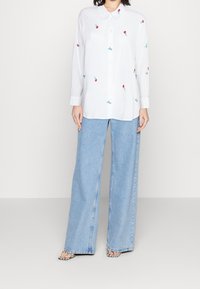 Chemise blanche à manches longues avec broderies florales et de bicyclette, associée à un jean denim bleu clair à jambes larges, portée avec des sandales à motifs.