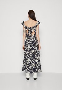 Robe maxi bleu marine avec motif floral blanc, manches volantées, dos nu avec détail à nouer, et tissu fluide. Associée à des bottes à talons blanches.