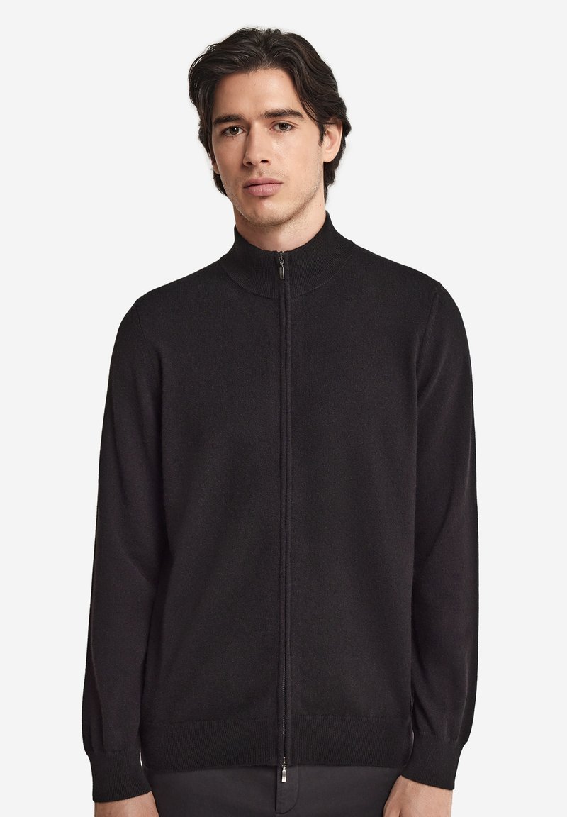 Junger Mann mit dunklen Haaren, der einen schwarzen Pullover mit durchgehendem Reißverschluss und hohem Kragen vor weißem Hintergrund trägt.