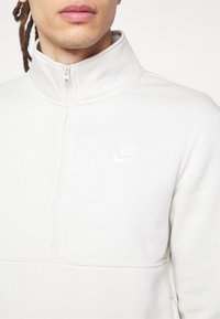 Jeune personne portant un sweat-shirt Nike blanc à demi-fermeture éclair, montrant le col, la fermeture éclair et le logo Nike brodé blanc sur la poitrine.