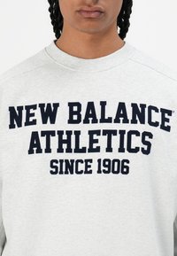 Persona ar gaišas pelēkas sporta virsjakas, kurai uz krūtīm ir melns uzraksts "NEW BALANCE ATHLETICS SINCE 1906".