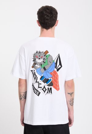 Homme portant un t-shirt blanc avec un graphisme coloré d’un personnage à tête de boule épineuse tenant une batte, assis sur un mur en briques, et texte stylisé.