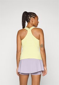 En person med flettehår står med ryggen til, iført en gul tanktop og en lilafarget skorts, som fremhever designet på aktivklær.