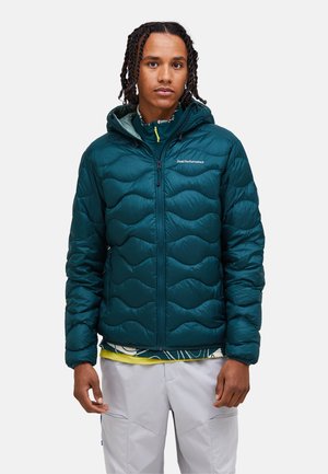 Ung mand med flettet hår iført en mørk teal quiltet hættetrøje og lysegrå bukser, stående mod en ensfarvet baggrund.