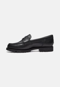 Mocassin en cuir noir à bout rond, bride décorative, détails cousus, et semelle en caoutchouc crantée pour une bonne adhérence, présenté en profil.