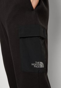 Pantalones de chándal negros de algodón con un gran bolsillo tipo carguero. Presentan un logo en relieve en un parche de tela. Textura suave con un ajuste relajado.