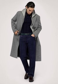 Šedý dlouhý kabát z vlny s připevněnou kapucí, doplněný o tmavě modré polo, tmavě modré kalhoty a hnědé kožené boty.