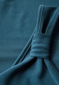 Tealfärgad tyg med en slät yta, som har en vriden knutdetalj och fint sydda kanter, visad i en närbild.